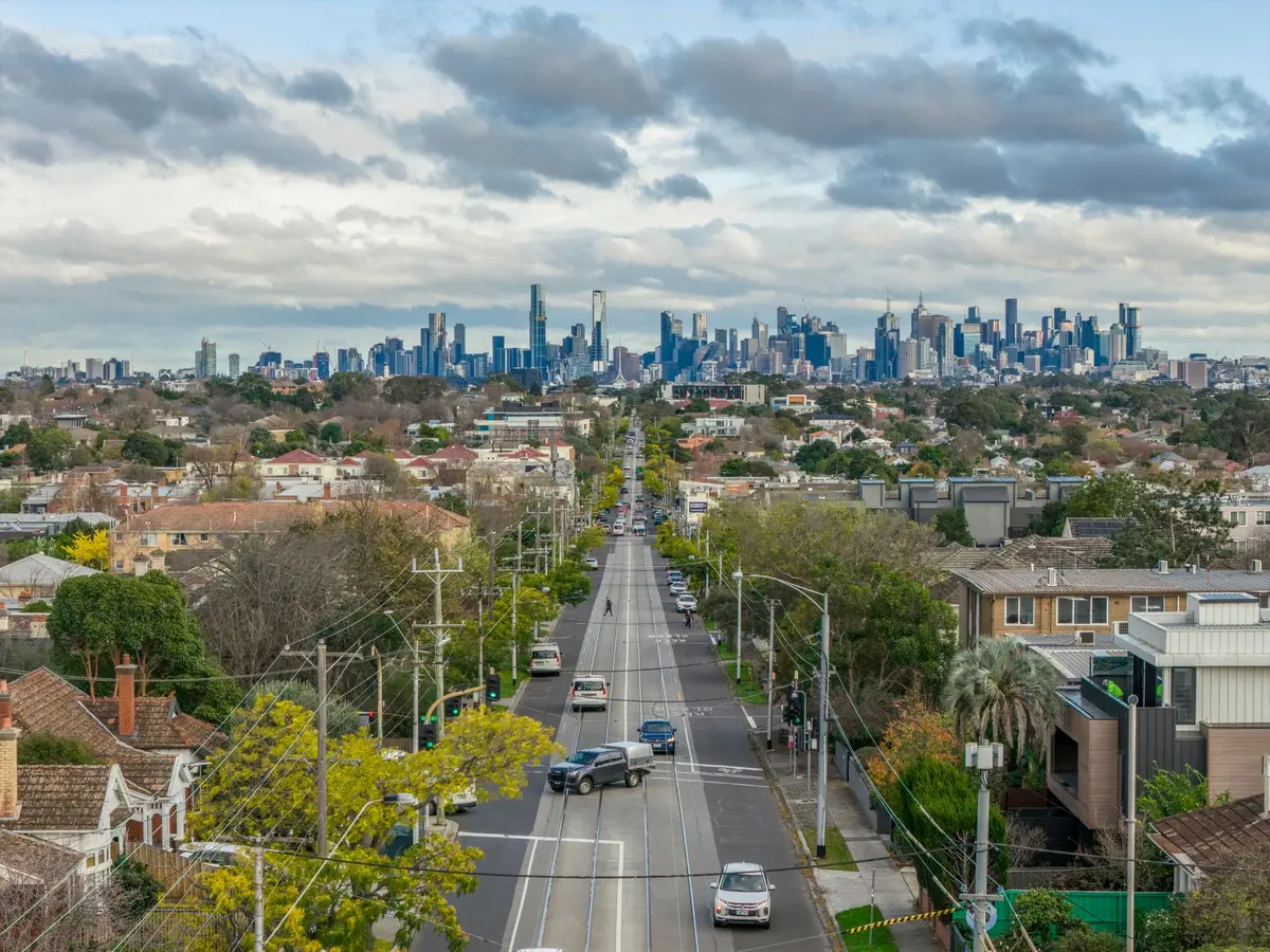 A tree-lined Melbourne street of terracotta-roofed houses running downhill toward the city skyline in the distance, tram wires overhead.